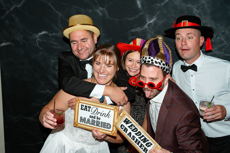 Wedding guests posing at an open-air photo booth with a floral backdrop and fun props