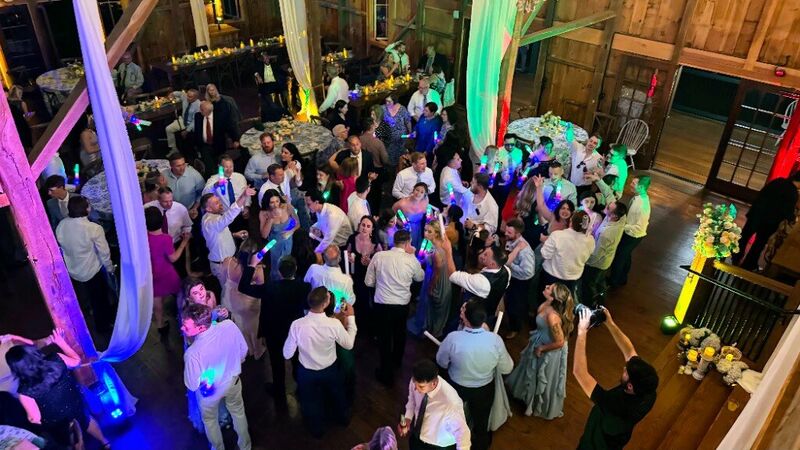 Elevated view of a packed barn wedding dance floor with guests waving glow sticks under colorful uplighting